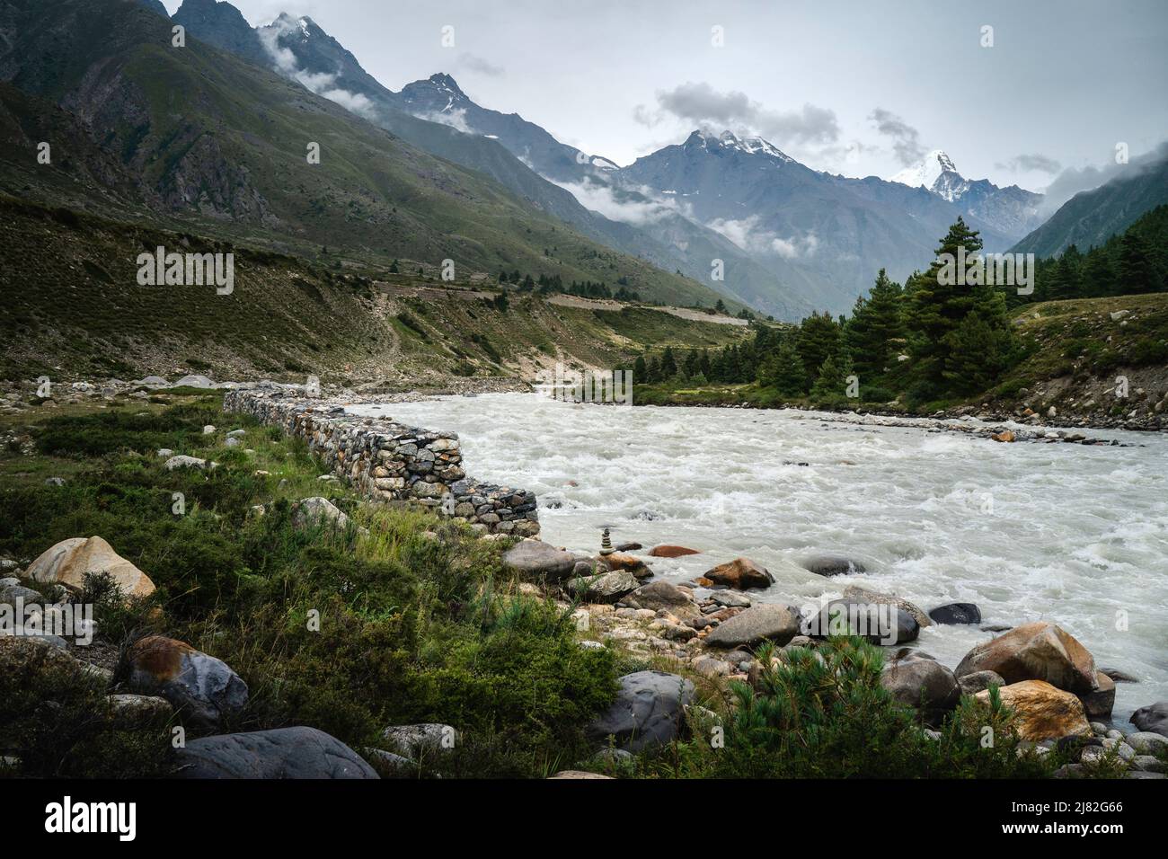 Baspa river flanked by boulders, trees, and the Himalayas with peaks shrouded in clouds under overcast sky in summer in Chitkul, India. Stock Photo
