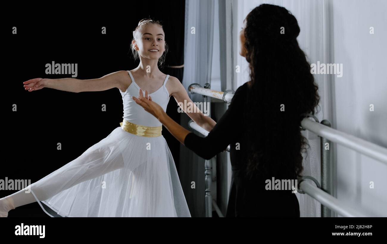 Caucasian girl teenager student ballerina in tutu doing dancing exercises near ballet barre listening to explanations advice from adult teacher female Stock Photo