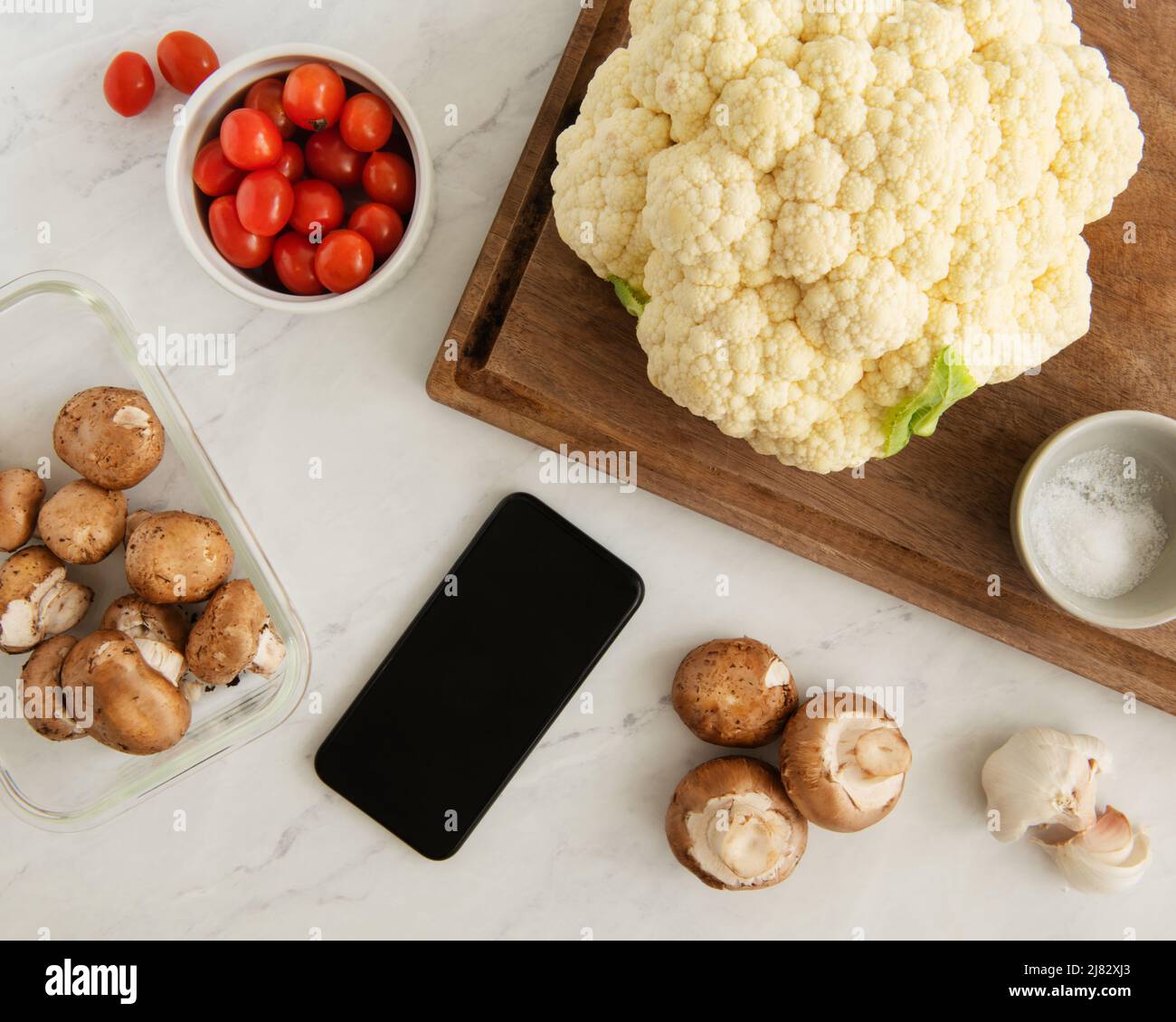 Cooking scene with Cauliflower, mushrooms, garlic, and tomatoes and smart phone for instructions Stock Photo