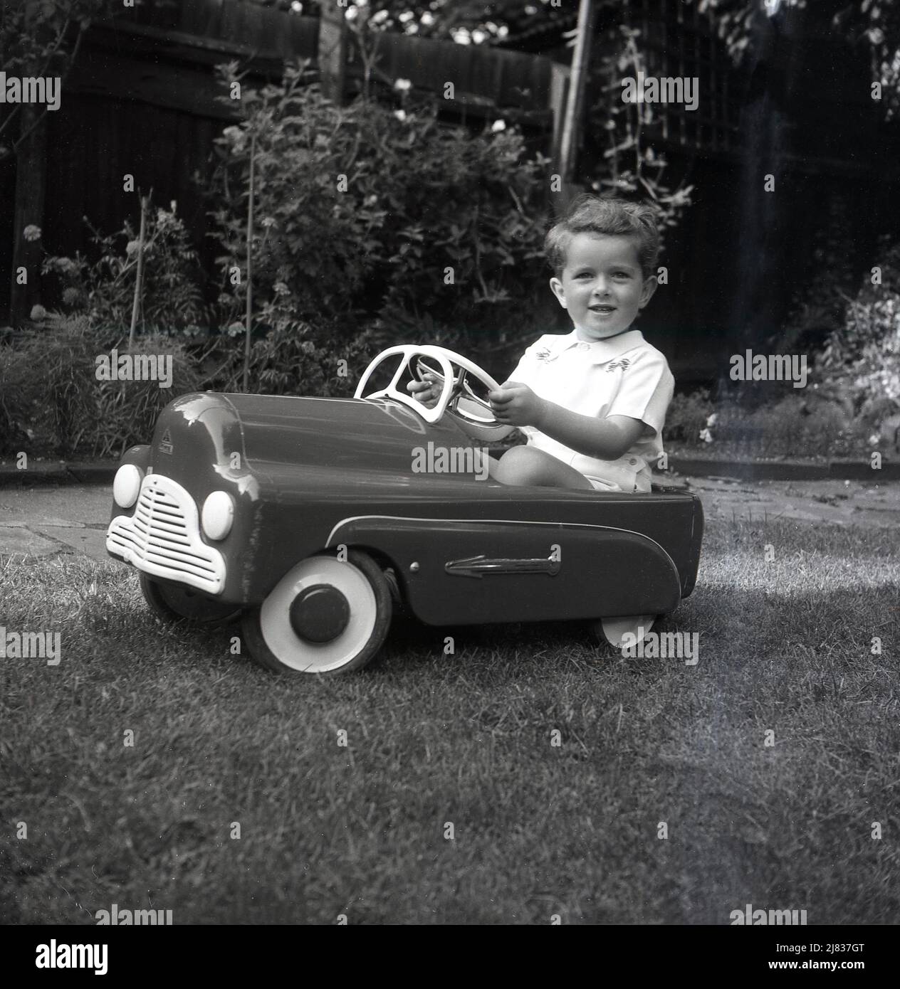 1953, historical, outside in a garden, a young boy sitting in his ride-on toy pedal car of the era, a British made Tri-Ang 'Thirty B'.  Made out of pressed steel, the toy car had a high gloss enamel finish, with Chrome arrows on the side. Tri-Ang was the brand name of Line Bros. Ltd, a British business, who in this era was the largest toy and baby carriage manufacturing company in the world. Stock Photo