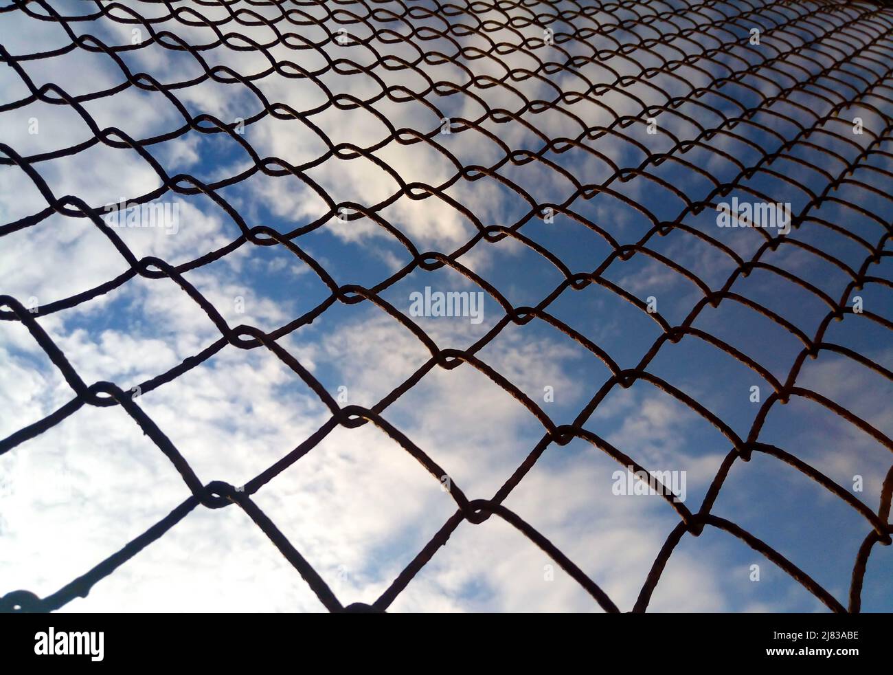 Bright blue sky with white clouds through a metal grid chain fence. Mesh background. Stock Photo