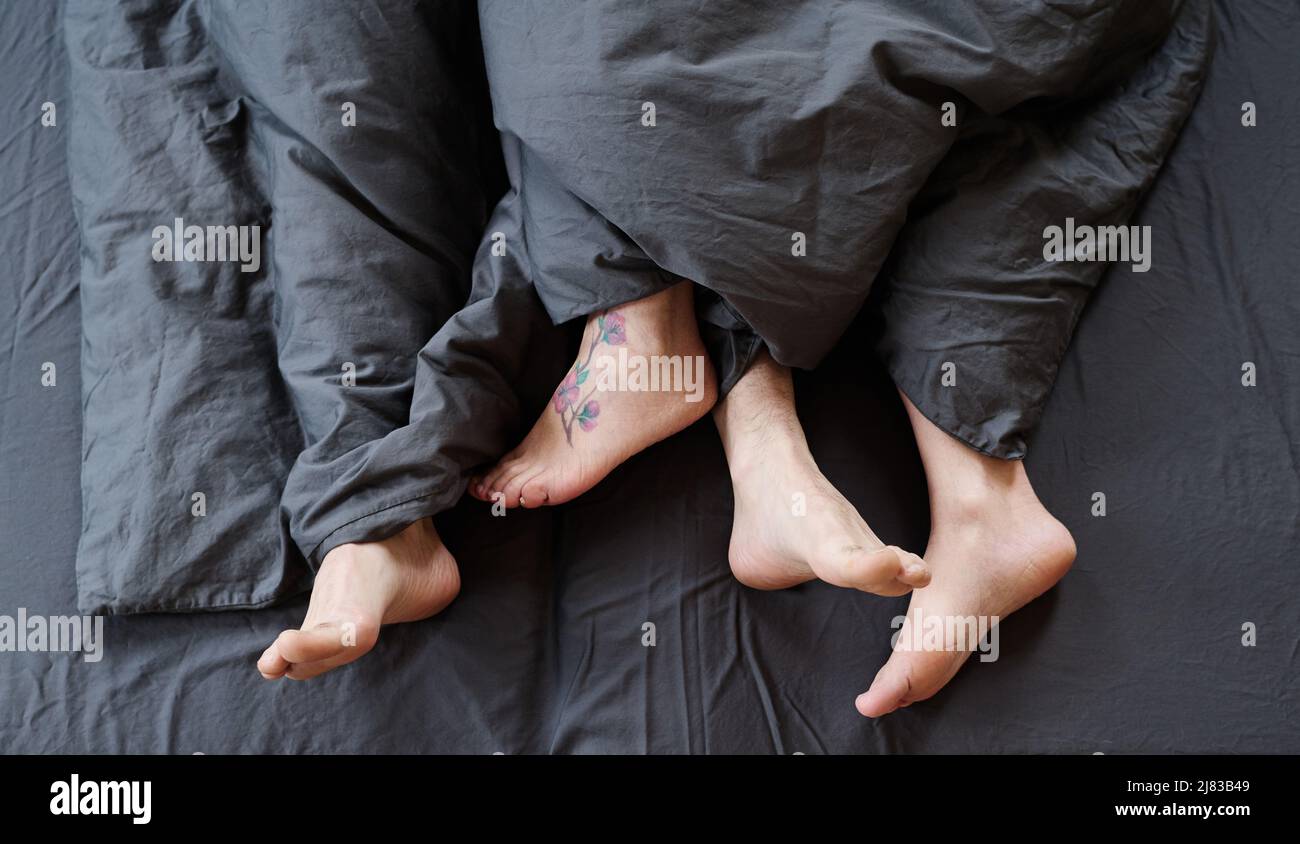 Top-down shot of modern unrecognizable romantic couple feet uncovered in bed in morning, gray bed linen Stock Photo