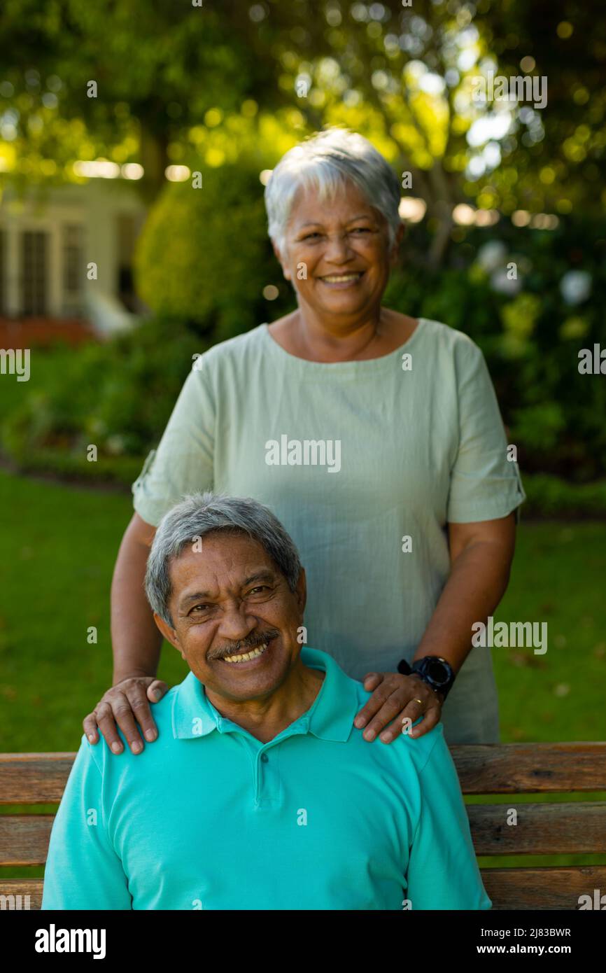 Portrait of smiling biracial senior woman standing behind senior husband sitting on bench in park Stock Photo