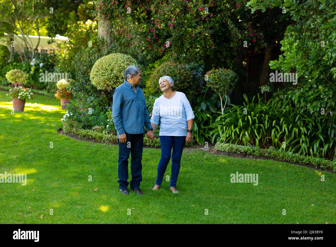 Biracial happy senior woman holding husband's hand while standing against plants and trees in park Stock Photo