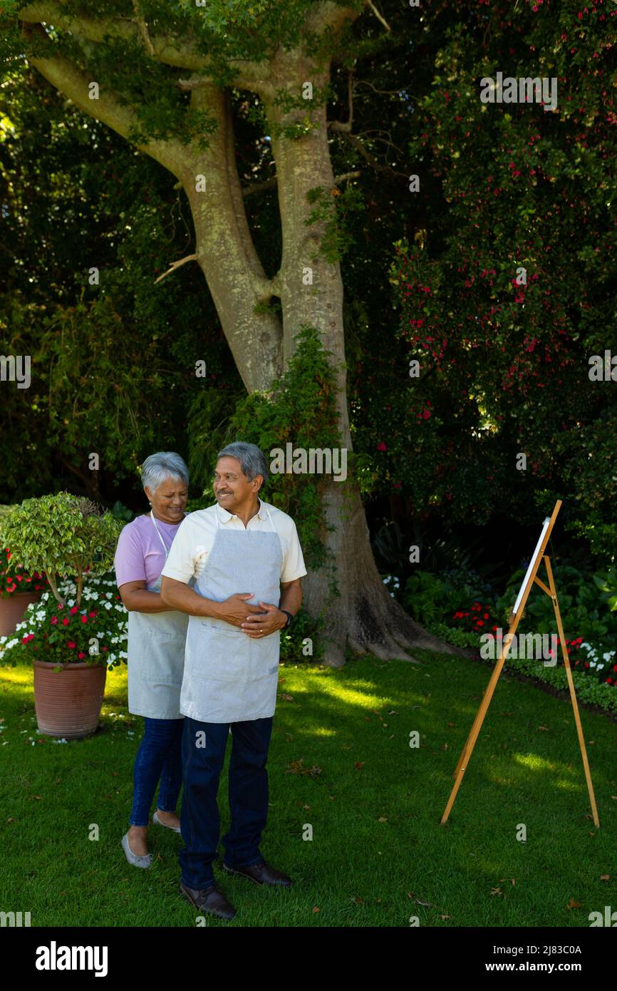 Biracial senior wife tying husband's apron standing by canvas and easel against plants in yard Stock Photo
