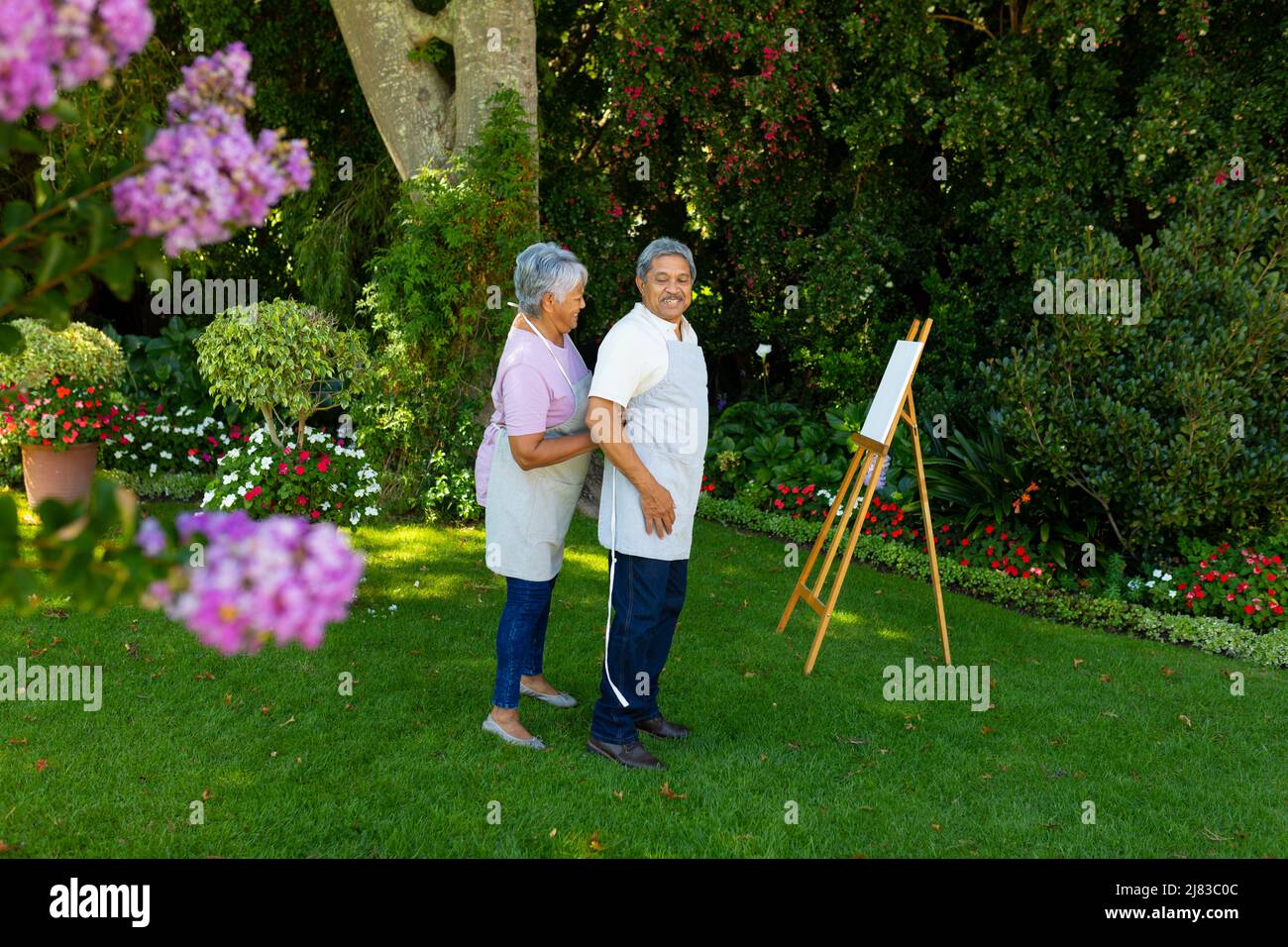 Biracial senior woman tying husband's apron standing by canvas and and easel against plants in yard Stock Photo