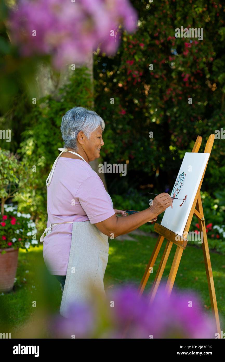 Side view of smiling biracial senior woman with short hair painting on canvas against plants in yard Stock Photo