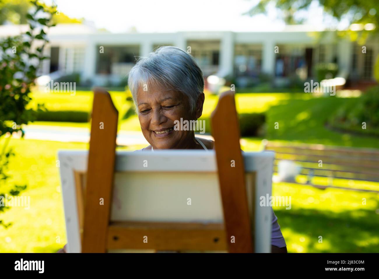 Smiling biracial senior woman with short gray hair painting on canvas against house in yard Stock Photo