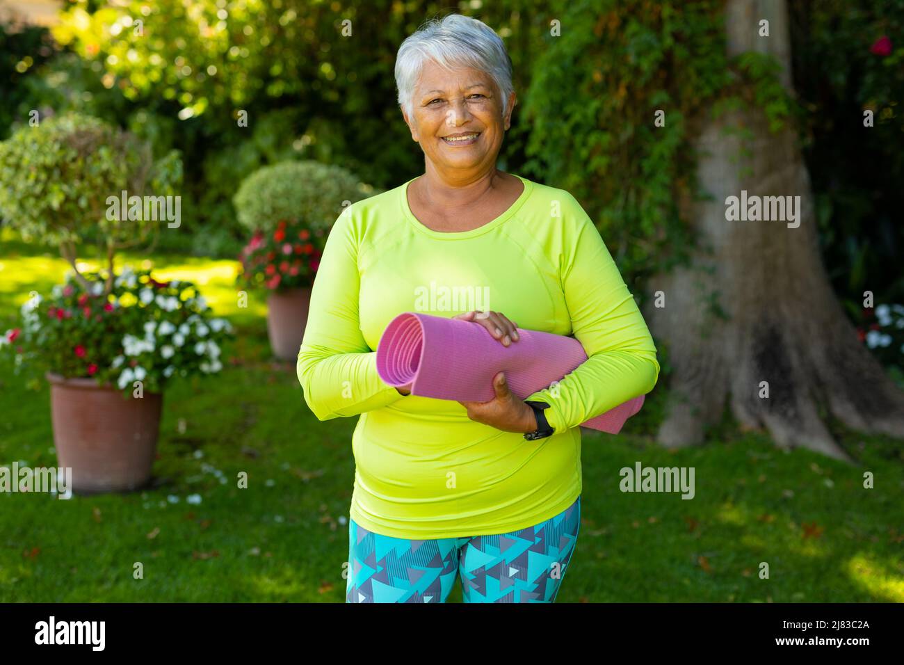 Portrait of smiling biracial senior woman wearing sports clothing holding yoga mat against plants Stock Photo