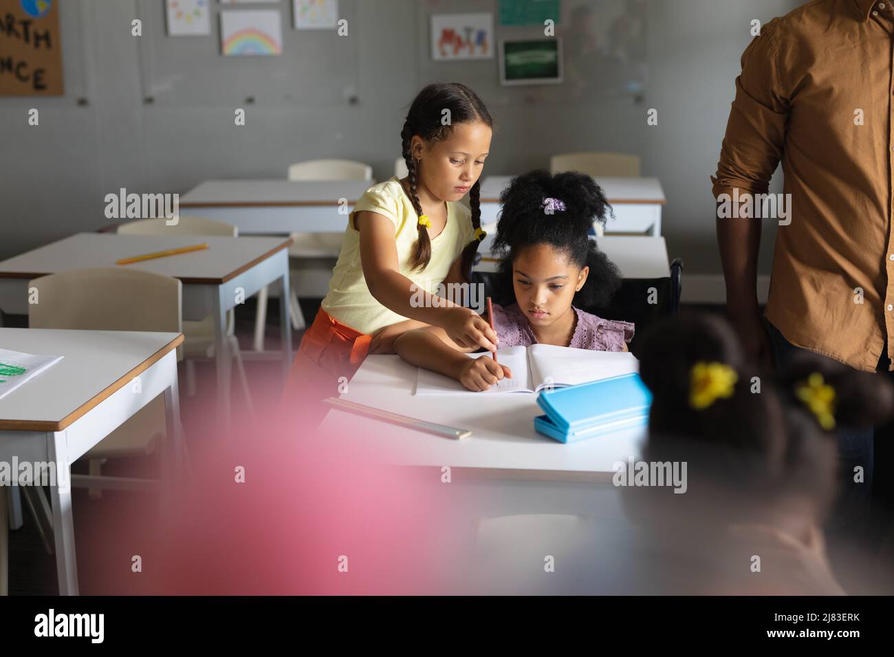 Caucasian elementary schoolgirl assisting biracial classmate studying at desk in classroom Stock Photo