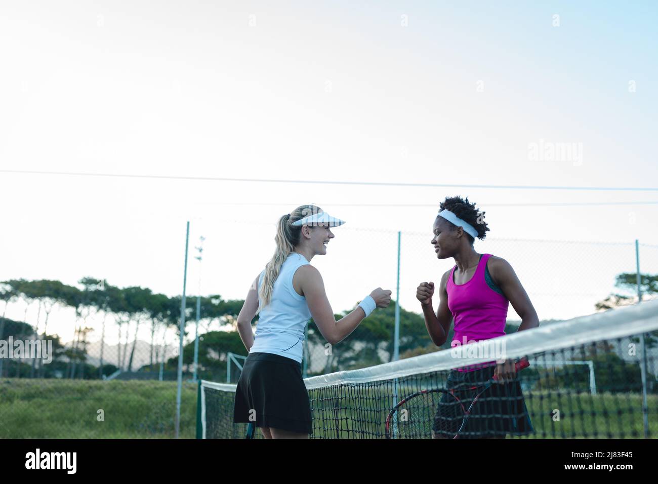 Happy multiracial young female players giving fist bump over net at tennis court Stock Photo