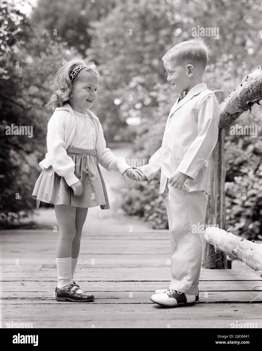 1960s SMALL BOY AND GIRL DRESSED UP STANDING ON WOODEN BRIDGE HOLDING HANDS SMILING AT ONE ANOTHER - j11319 HAR001 HARS JUVENILE STYLE COMMUNICATION FRIEND STRONG PLEASED JOY LIFESTYLE FEMALES HEALTHINESS HOME LIFE COPY SPACE FRIENDSHIP FULL-LENGTH PERSONS INSPIRATION BOYFRIEND CARING MALES CONFIDENCE B&W SUMMERTIME GIRLFRIEND SUIT AND TIE HAPPINESS CHEERFUL AND CHOICE SADDLE SHOES HOLDING HANDS UP RELATIONSHIPS SMILES SADDLE OXFORDS SHORT DRESS FRIENDLY JOYFUL STYLISH SUNDAY BEST PERSONAL ATTACHMENT PUPPY LOVE AFFECTION ANOTHER CARDIGAN EMOTION JUVENILES MARY JANE SHOES SEASON TOGETHERNESS Stock Photo