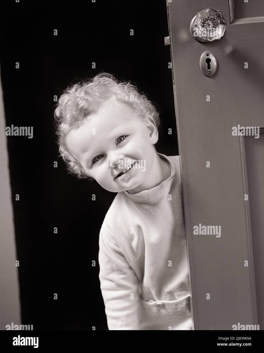 1930s 1940s SMILING LITTLE GIRL WITH CURLY HAIR PEEKING AROUND A DOOR - j2599 HAR001 HARS SATISFACTION FEMALES WINNING STUDIO SHOT HEALTHINESS HOME LIFE COPY SPACE FRIENDSHIP HALF-LENGTH PAJAMAS CONFIDENCE B&W EYE CONTACT HAPPINESS CHEERFUL ADVENTURE DISCOVERY CURLY SMILES JOYFUL PLEASANT AGREEABLE CHARMING GROWTH JUVENILES LOVABLE PLEASING ADORABLE APPEALING BABY GIRL BLACK AND WHITE CAUCASIAN ETHNICITY HAR001 OLD FASHIONED Stock Photo