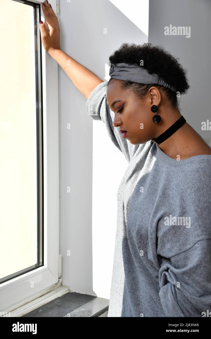 Eine nachdenkliche Afro-Amerikanische junge huebsche Frau steht an einem Fenster und blickt nachdenklich zu Boden. Sie traegt einen grauen Pullover Stock Photo