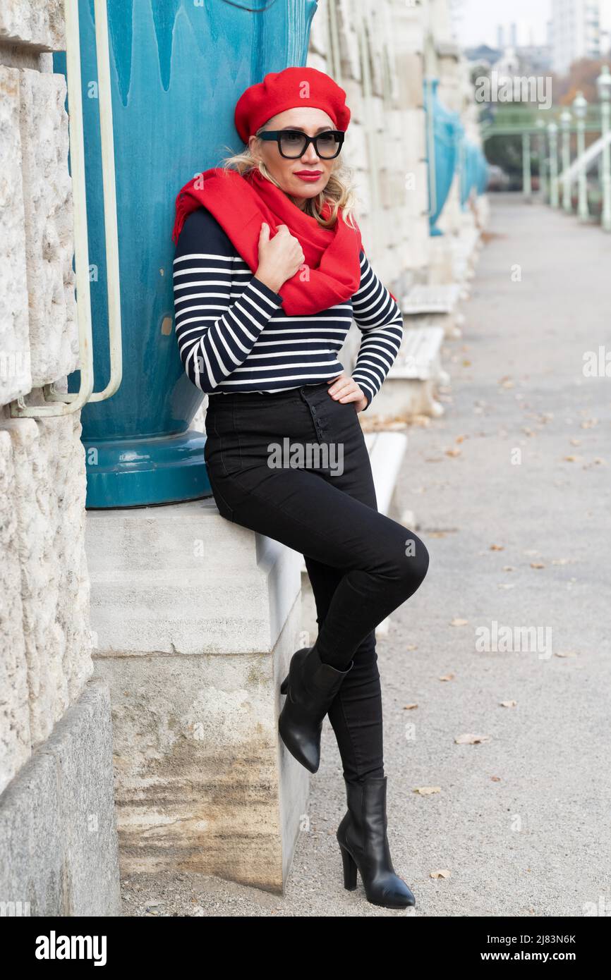 In einem Park lehnt an einer Mauer eine gut aussehende Dame mit roter Muetze und passendem Schal. Die schwarze Hose mit den Stiefeln passt zum Stock Photo