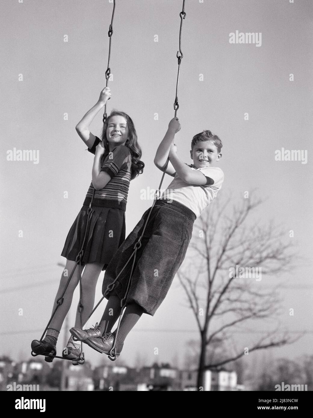 1930s SMILING PRETEEN BOY AND GIRL STANDING ON PLAYGROUND PARK CHAIN SWING SEAT LOOKING AT CAMERA - j7767 HAR001 HARS HEALTHY FRIEND BALANCE SAFETY TEAMWORK PANTS PLEASED JOY LIFESTYLE FEMALES RURAL CHAIN COPY SPACE FRIENDSHIP FULL-LENGTH PERSONS TRADITIONAL MALES RISK CONFIDENCE B&W EYE CONTACT ACTIVITY HAPPINESS PHYSICAL CHEERFUL STRENGTH TROUSERS AND CHOICE LOW ANGLE RECREATION PRIDE ON PRETEEN SMILES SWINGING KNEE SOCKS CLINGING CONCEPTUAL FLEXIBILITY FRIENDLY JOYFUL MUSCLES STYLISH SWINGS KNICKERBOCKERS PLUS FOURS GROWTH JUVENILES PRE-TEEN PRE-TEEN BOY PRE-TEEN GIRL TOGETHERNESS Stock Photo