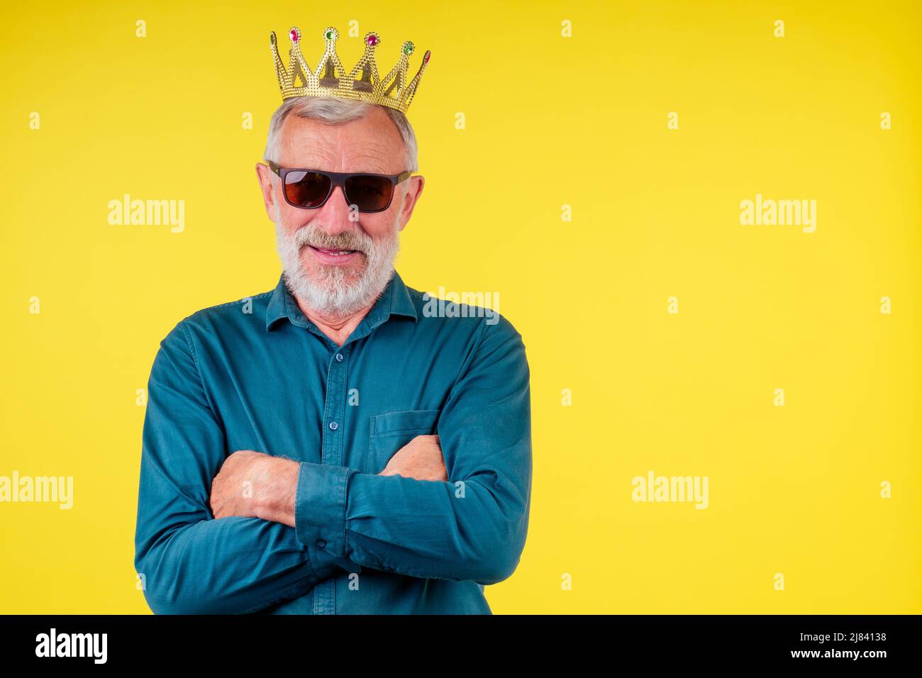 close-up portrait of happy senior man looking at camera , wearing green cotton shirt with a collar,gold crown and sunglasses in studio yellow Stock Photo