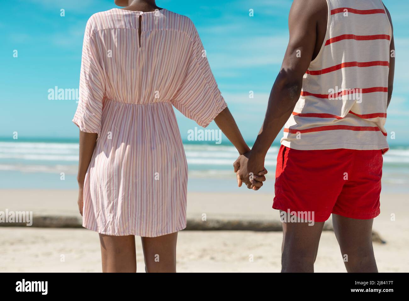 Midsection of african american young couple holding hands while standing at beach against blue sky Stock Photo