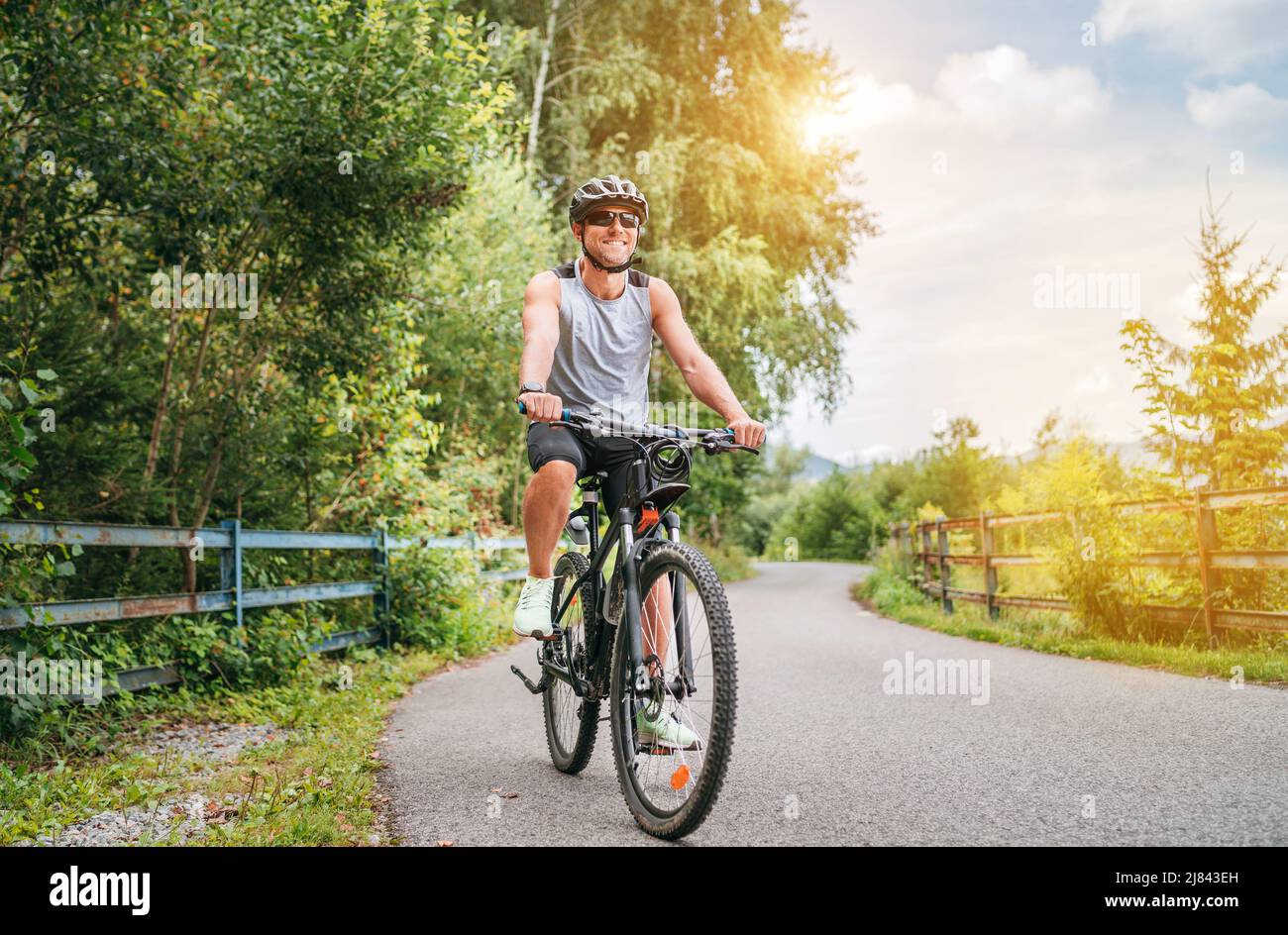 Portrait of a happy smiling man dressed in cycling clothes, helmet and sunglasses riding a bicycle on the asphalt out-of-town bicycle path. Active spo Stock Photo