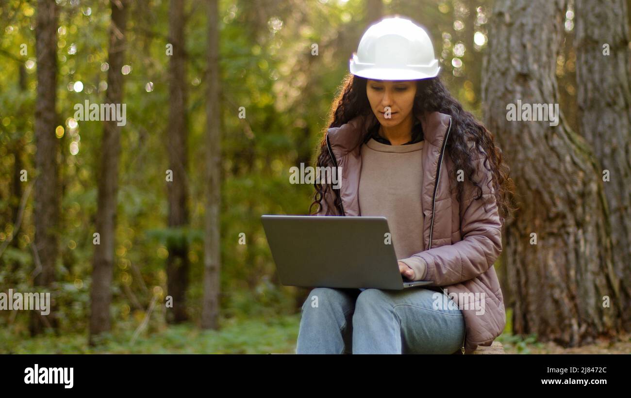 Millennial woman young experienced specialist forestry engineer environmentalist technician in hardhat checking trees entering data into laptop taking Stock Photo