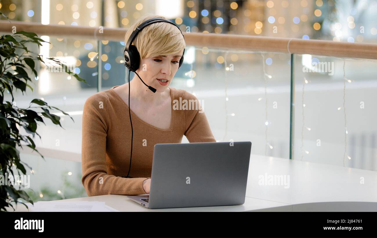 Middle aged adult 40s business woman in headset microphone talking at laptop web camera consultation online distant remote teacher coach helpline Stock Photo