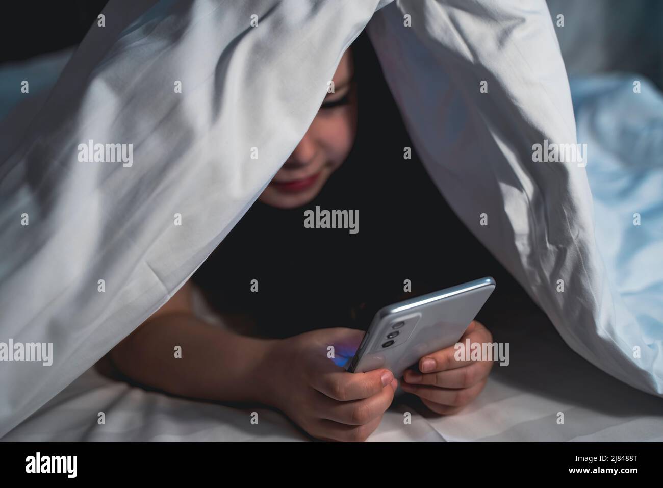 cute little girl hides under blanket with smartphone at night when
