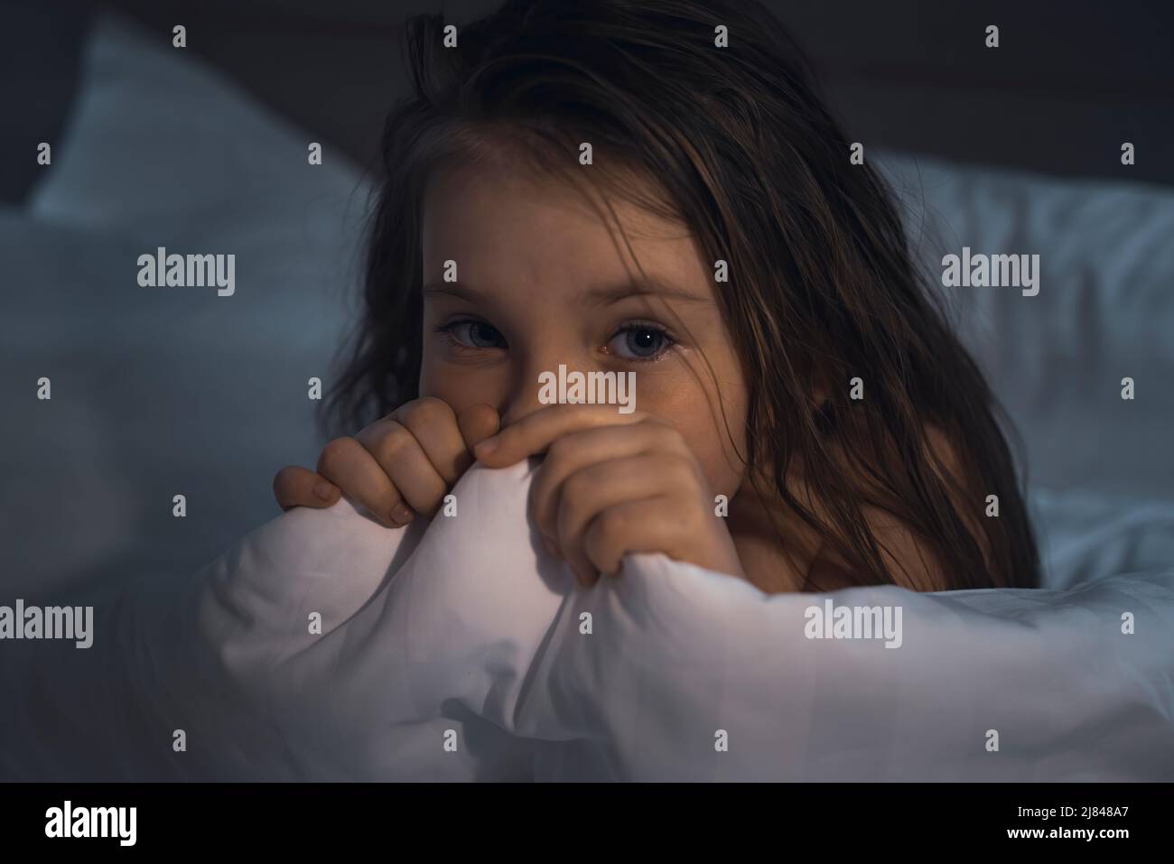 A cute little girl lies in bed, frightened and covers her face with a blanket. children. High quality photo Stock Photo