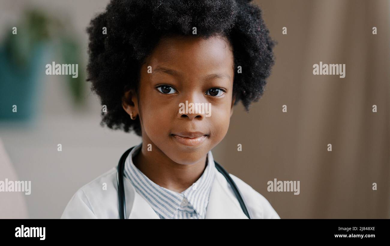 Cute serious african american kid girl in medical clothes dressed in white coat standing indoors posing looking at camera playing pretending to be Stock Photo