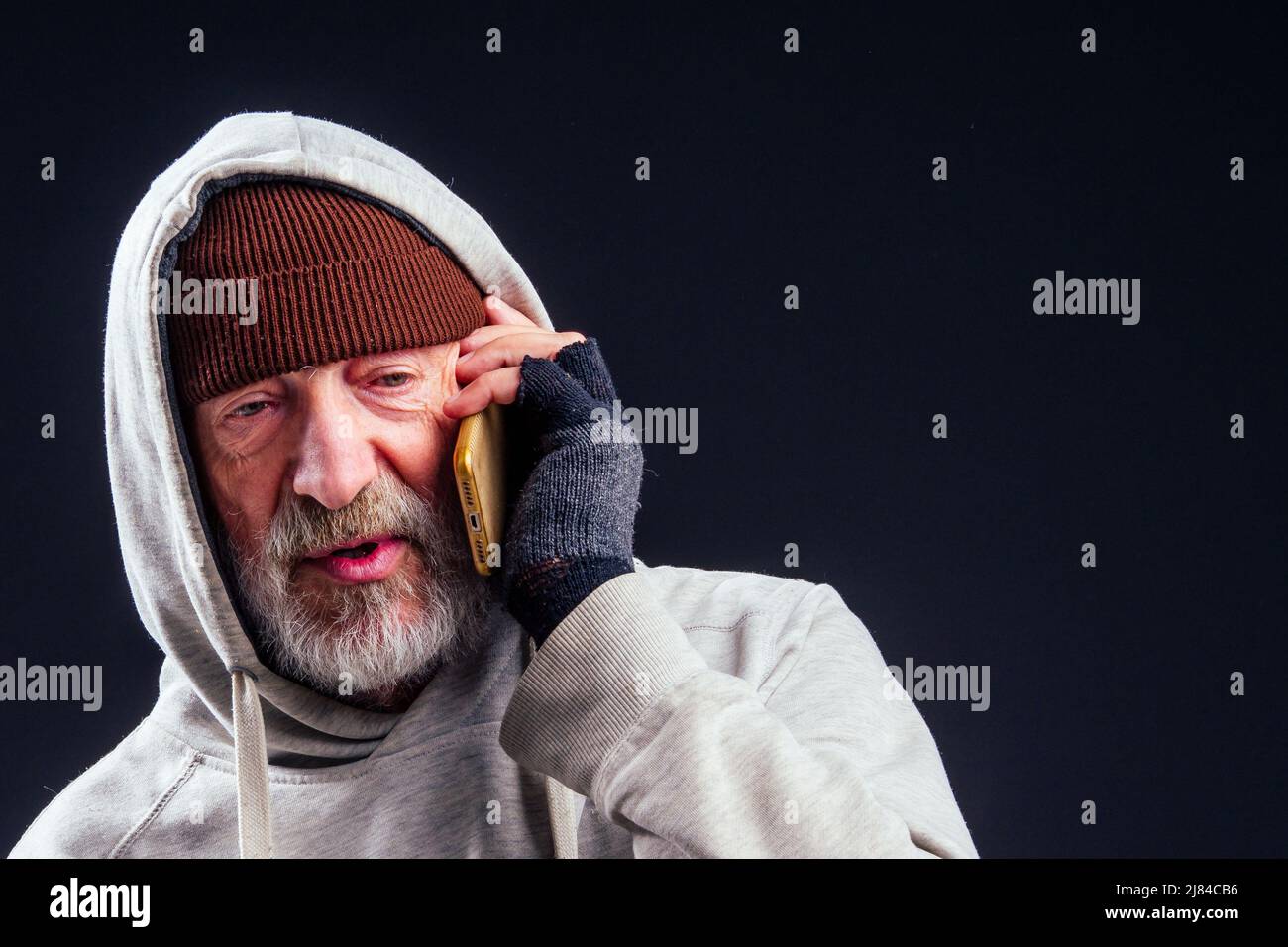 portrait of sad old senior male tramp wearing warm brown hat and leaky gloves,talking by smartphone in studio black background Stock Photo