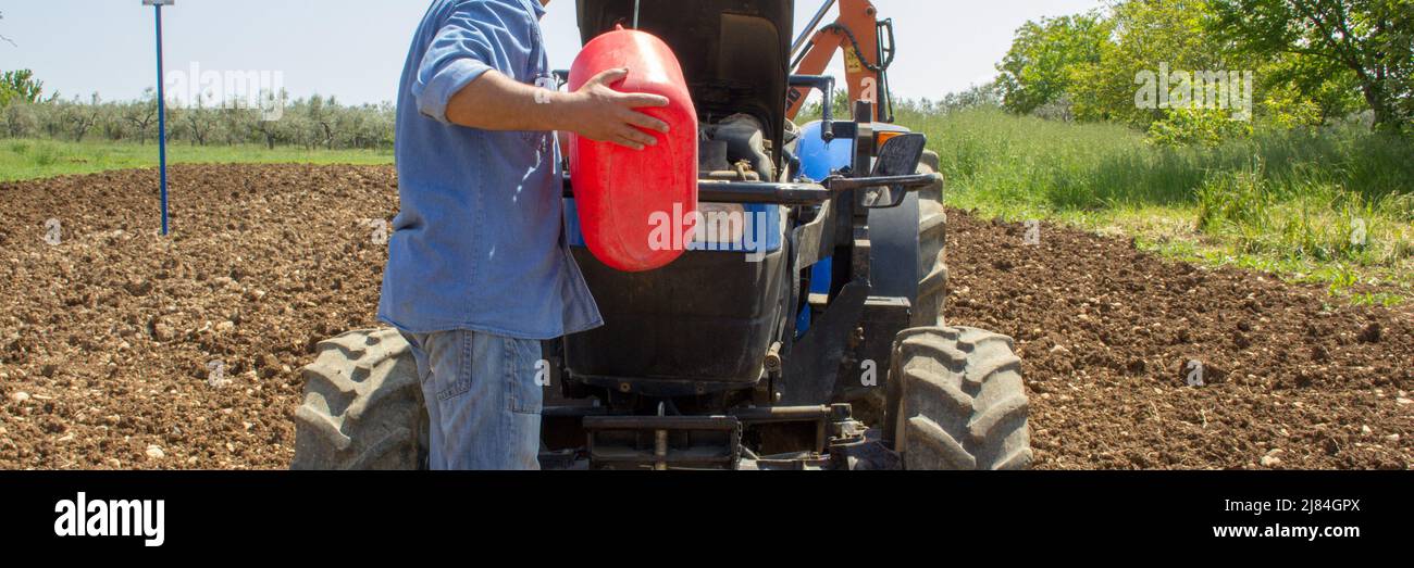 Farmer refueling his tractor with a jerry can, as he runs out of fuel. Reference to the expensive and current gasoline crisis. Horizontal banner Stock Photo