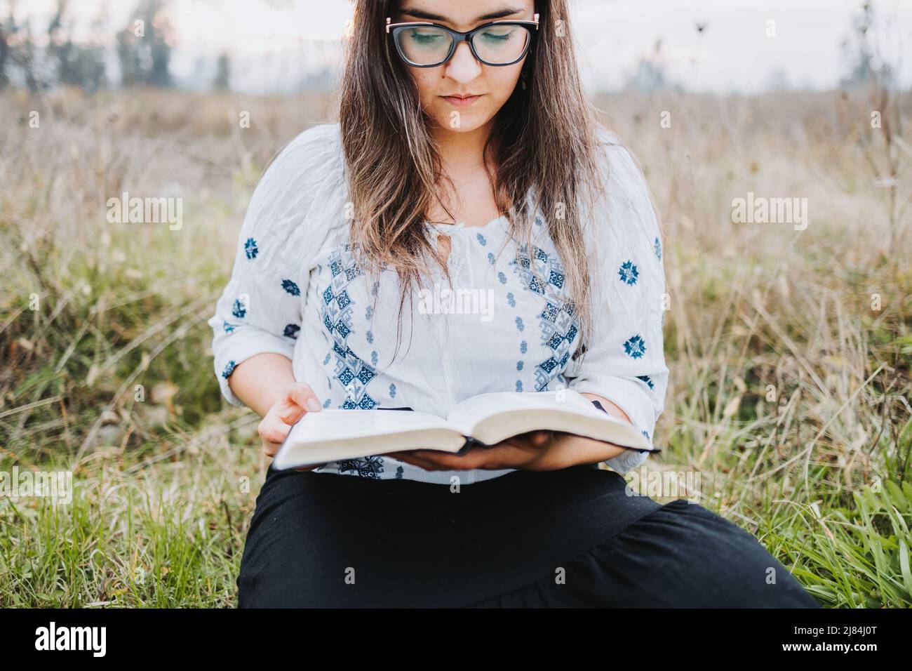 Woman with glasses sitting outdoor on the grass holding and reading an open Bible. Selective focus. Stock Photo