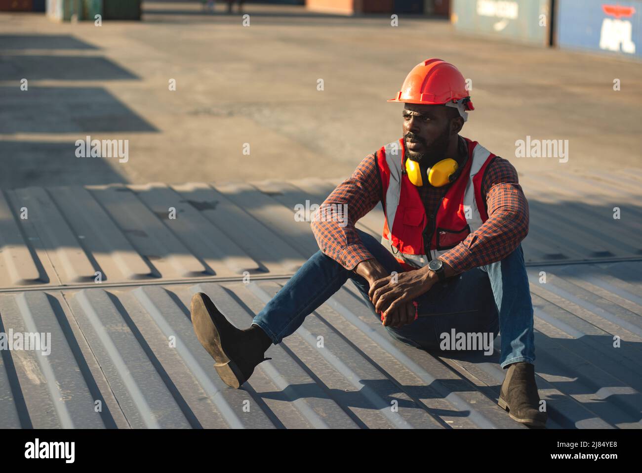 African American male technician or engineer. Sit near a container and look tired and sleepy or unemployed. Logistics industrial cargo mover concept. Stock Photo