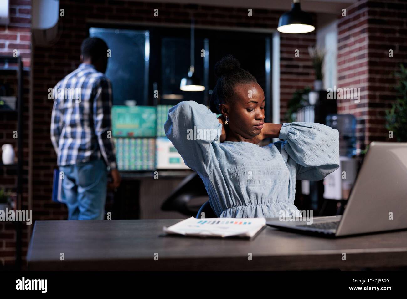 Tired financial analyst with neckache suffering from overwork hours. Forex stock market trading agency professional agent with neck pain sitting at desk in office workspace after a long day of work. Stock Photo