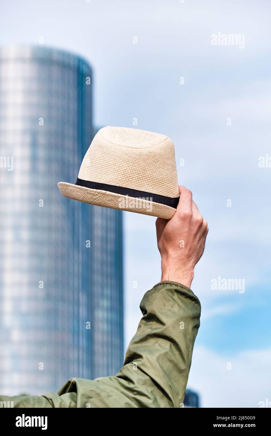 Man's hat in hand against the backdrop of office building. Welcome and greeting hand gesture, goodbye office and freedom concept. Stock Photo