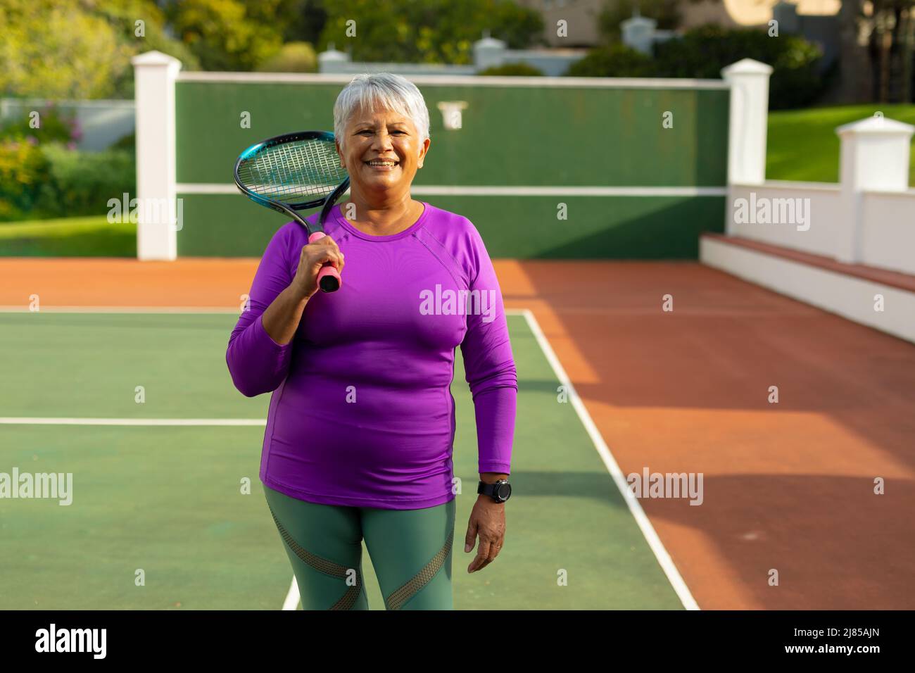 Portrait of confident smiling biracial senior woman with short hair holding tennis racket in court Stock Photo