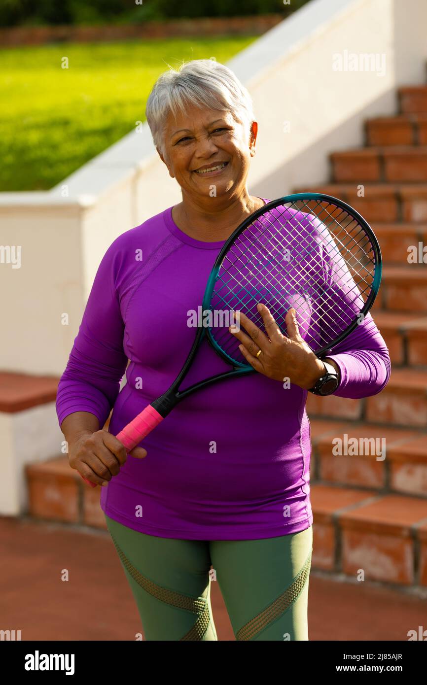 Portrait of smiling biracial senior woman with short hair holding tennis racket against steps Stock Photo