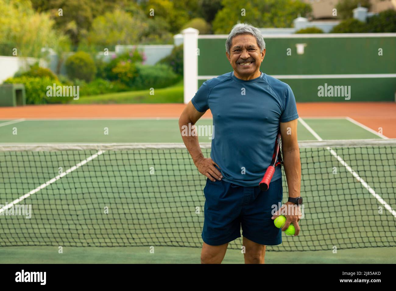 Portrait of happy biracial senior man with hand on hip holding balls and racket at tennis court Stock Photo