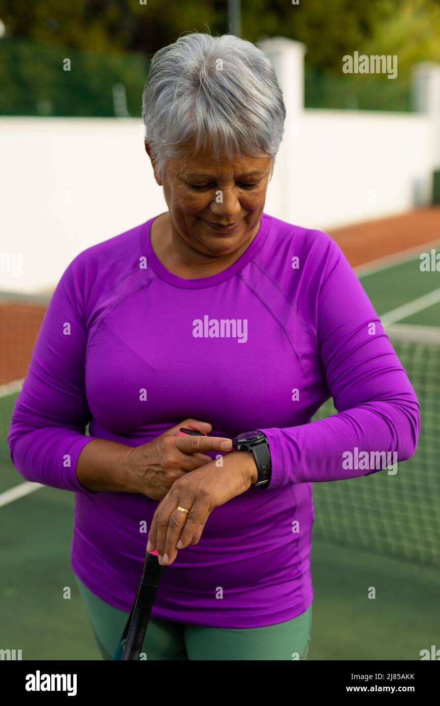 Smiling biracial senior woman with short hair holding tennis racket and checking time on wristwatch Stock Photo