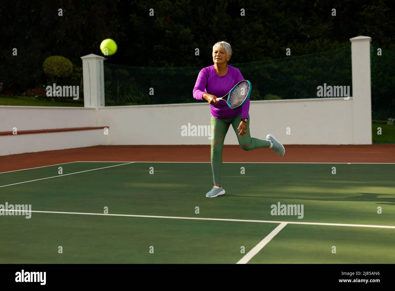 Biracial senior woman with short hair hitting tennis ball with racket while playing tennis at court Stock Photo