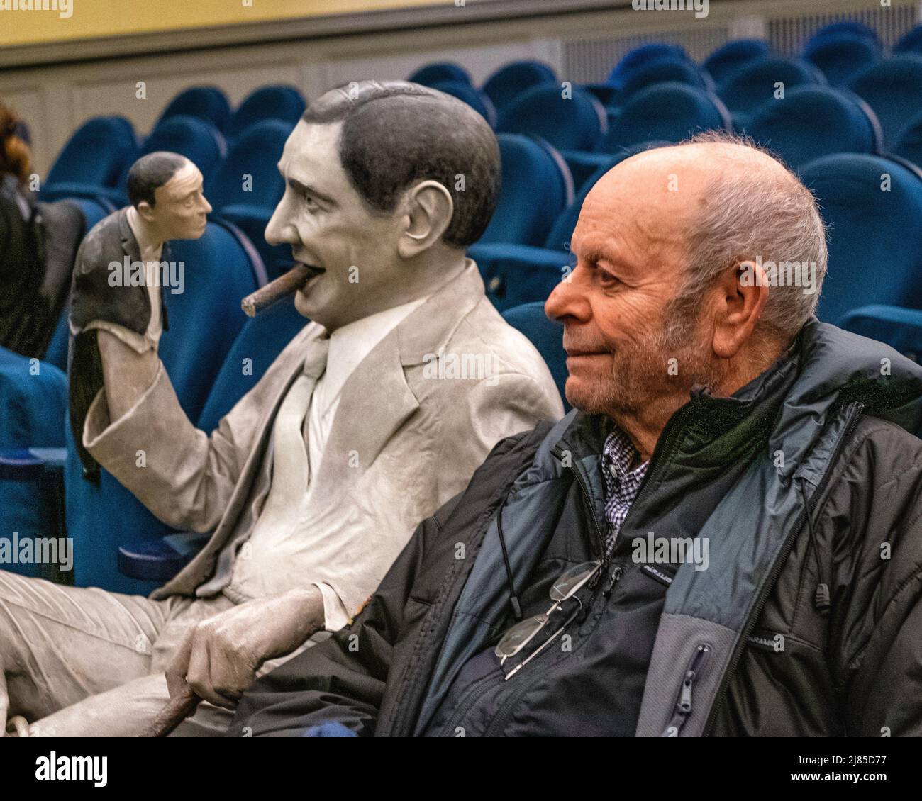 Senior elderly man sitting next to Statue of filmmaker Ernst Lubitsch, smoking a cigar, seated inside the movie theatre, Mitte, Berlin, Germany The statue sat in Schöneberg’s Notausgang cinema in the nineties and after the cinema closed down, I was moved to the Museum for Film and Television in the Sony Center. It has now been returned to a movie theatre. The Kino Babylon opened 1929 shows vintage films often with a with live orchestra Stock Photo