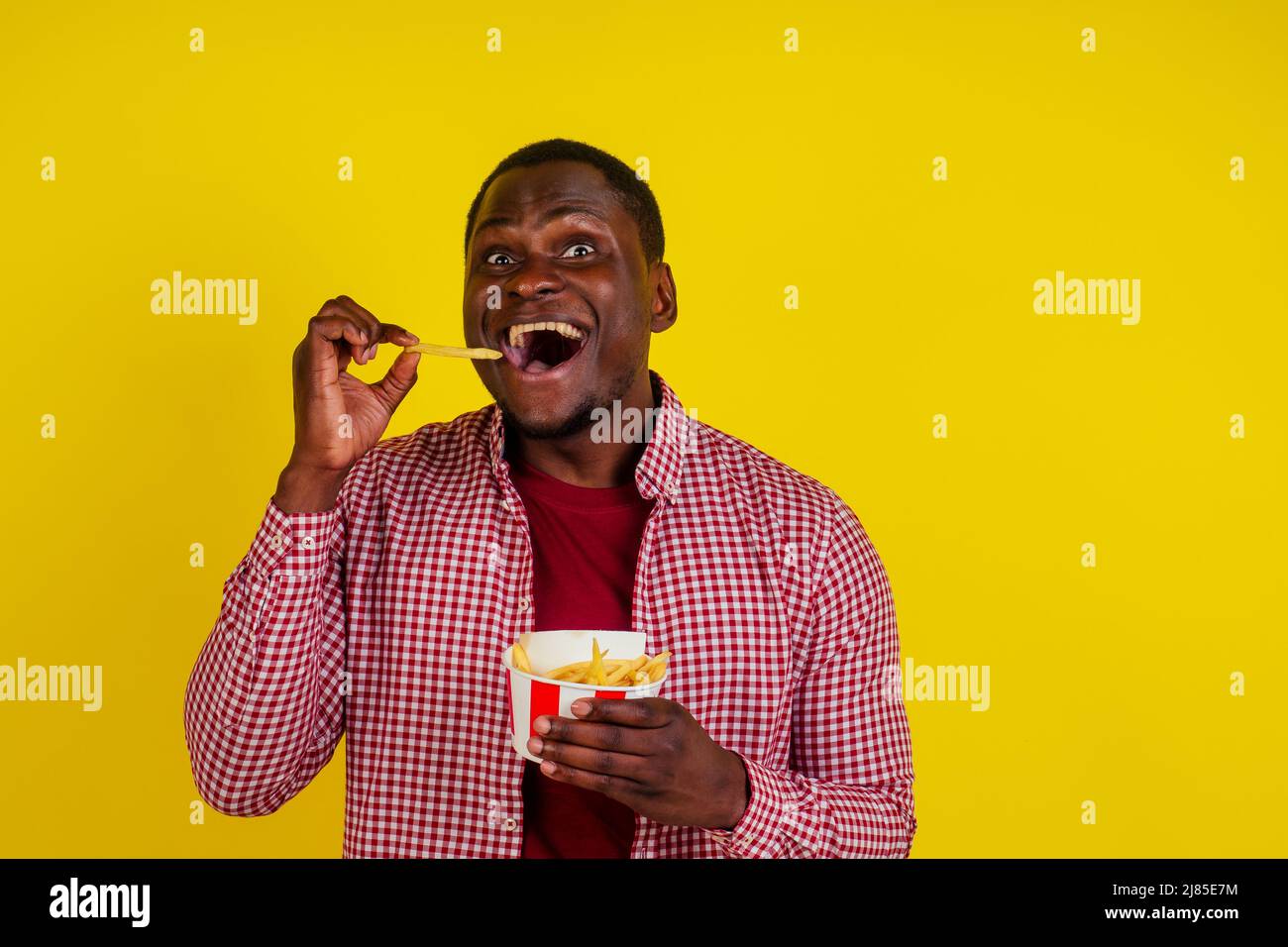 handsome latin man eating fries with appetite and enjoyment in studio yellow background Stock Photo