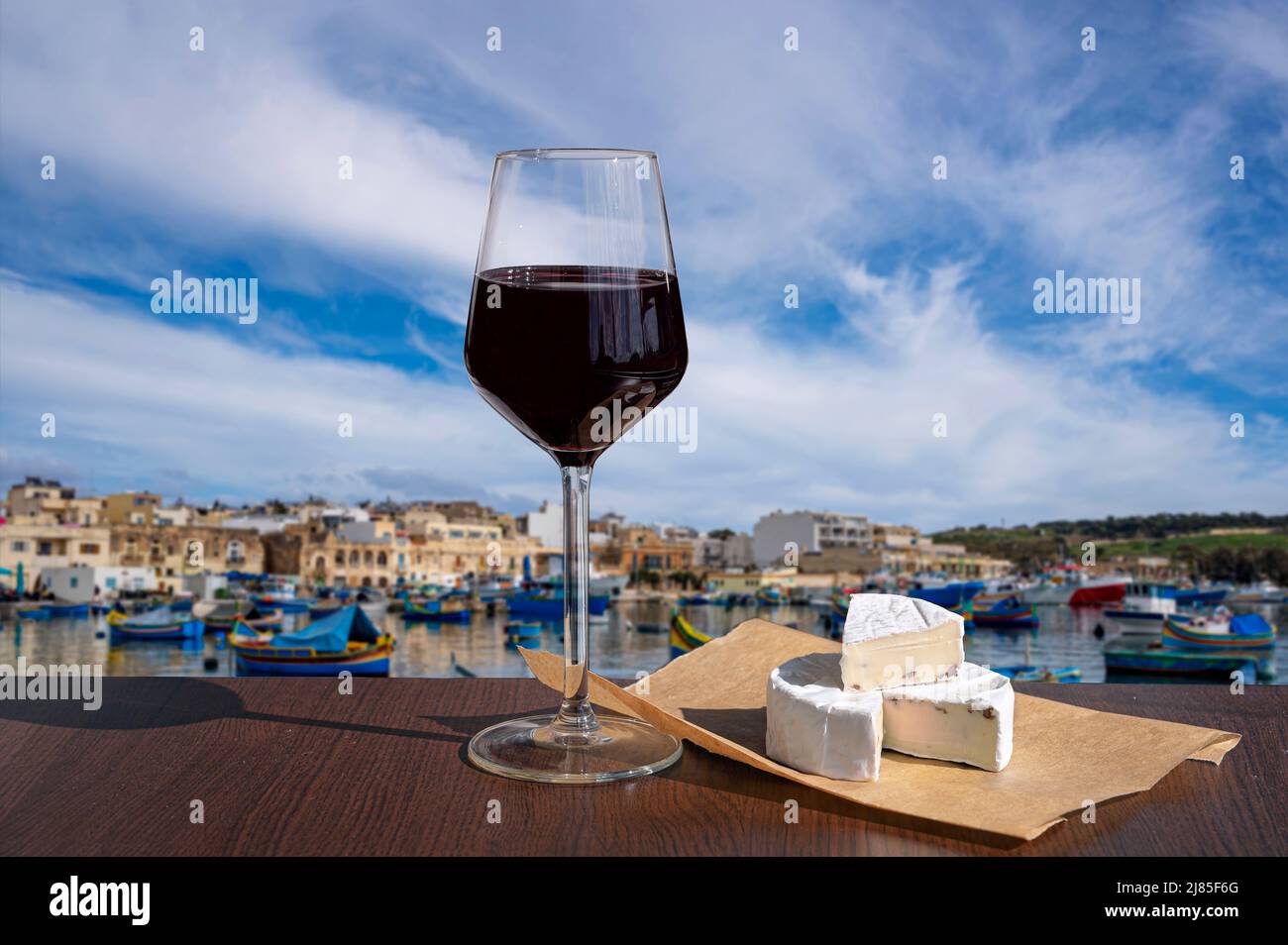 Glass of red wine with brie cheese with view of harbor with traditional boats and historic city center in Marsaxlokk, Malta Stock Photo