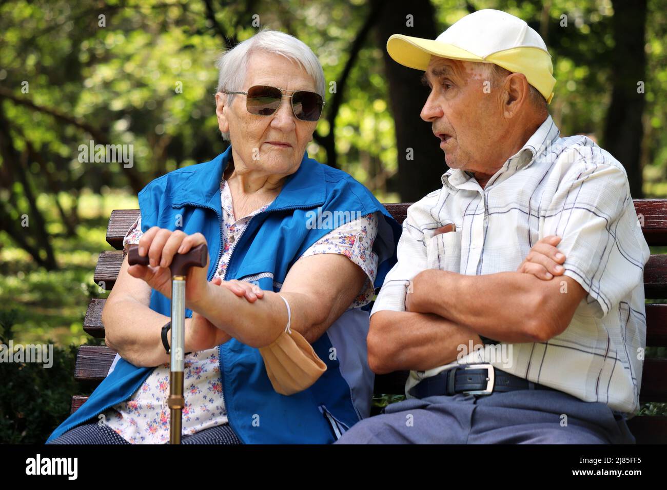 Elderly couple sitting on a bench in summer park. Old man and woman talking outdoors, life in retirement Stock Photo