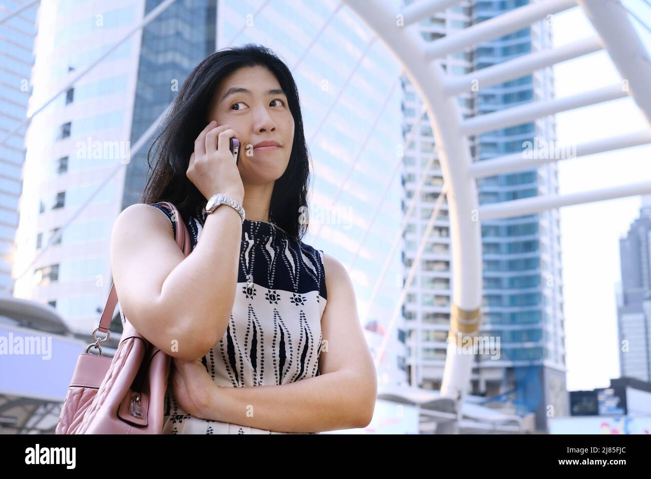 A young attractive Asian business woman in white dress is making a face while talking on her phone, feeling doubtful and skeptical. Stock Photo