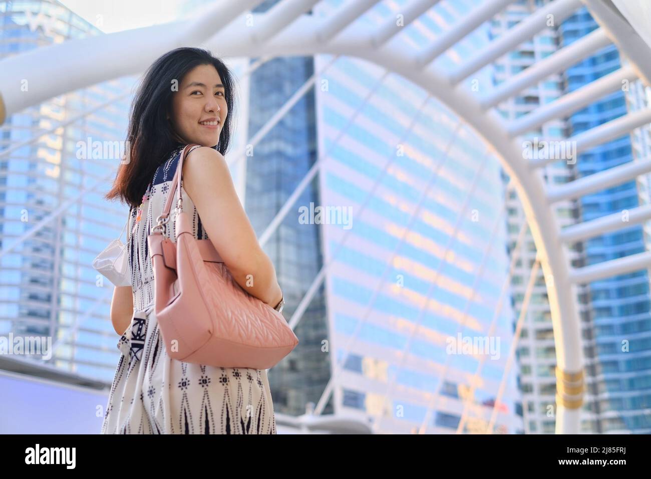 The back view of a young business woman walking on an intersection overpass in downtown Bangkok with high-rise office buildings in the background. Stock Photo