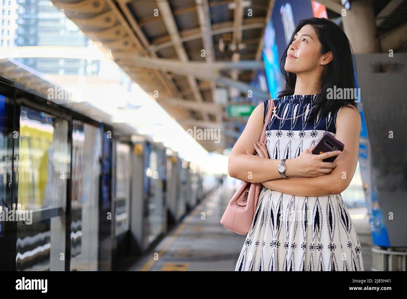 An Asian businesswoman in white and blue dress is standing with her arms crossed, waiting for the sky train at the station to go to work in the mornin Stock Photo