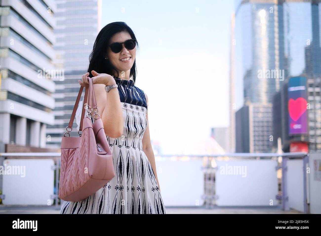 A beautiful confident Asian businesswoman in white and blue dress is holding her pink handbag, standing on an overpass walkway with the city skyline i Stock Photo