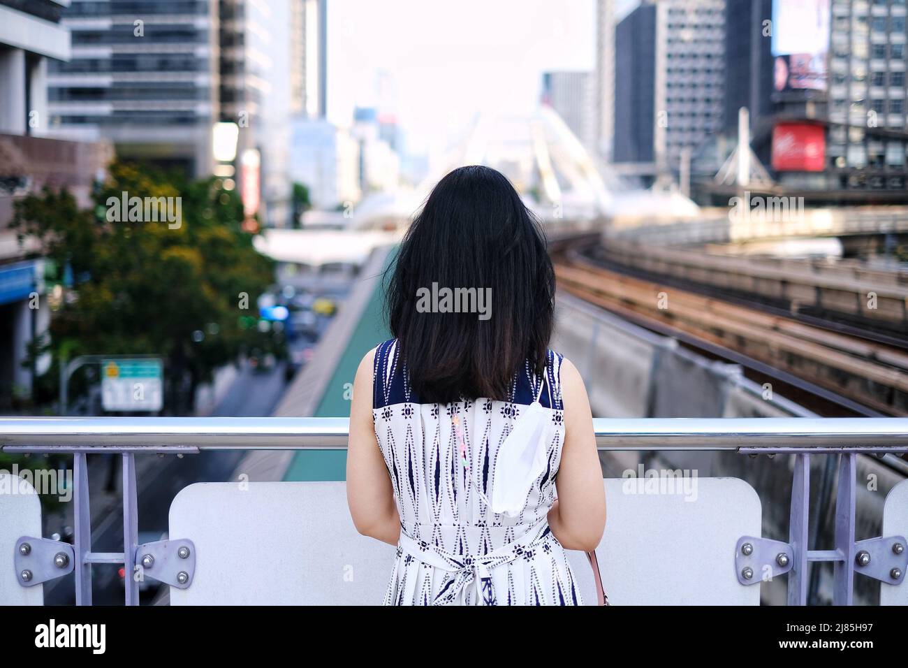 The back view of a woman in white and blue dress is standing on a walkway at a sky train station looking over the street and skyline of Bangkok Stock Photo