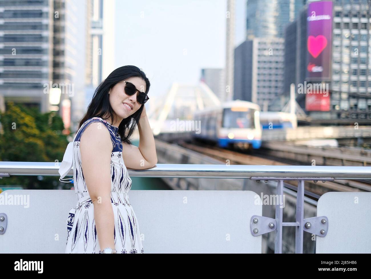 An Asian businesswoman is standing next to a walkway at a sky train station with the view of the incoming train on a track and the city skyline. Stock Photo
