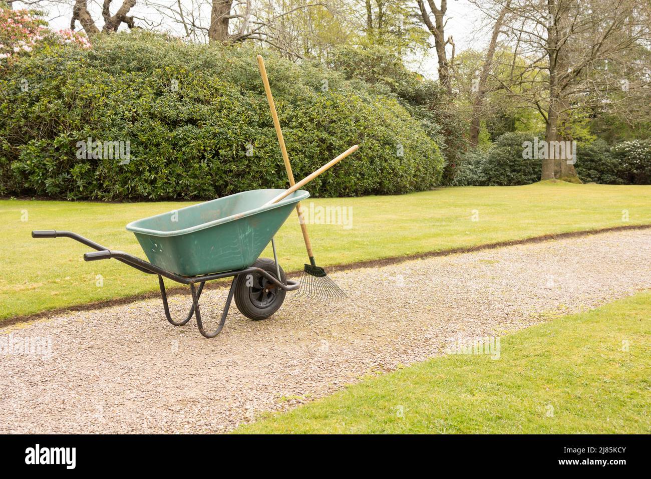 gardeners wheel barrow in green and tools on a gravel path and a well kept lawn Stock Photo